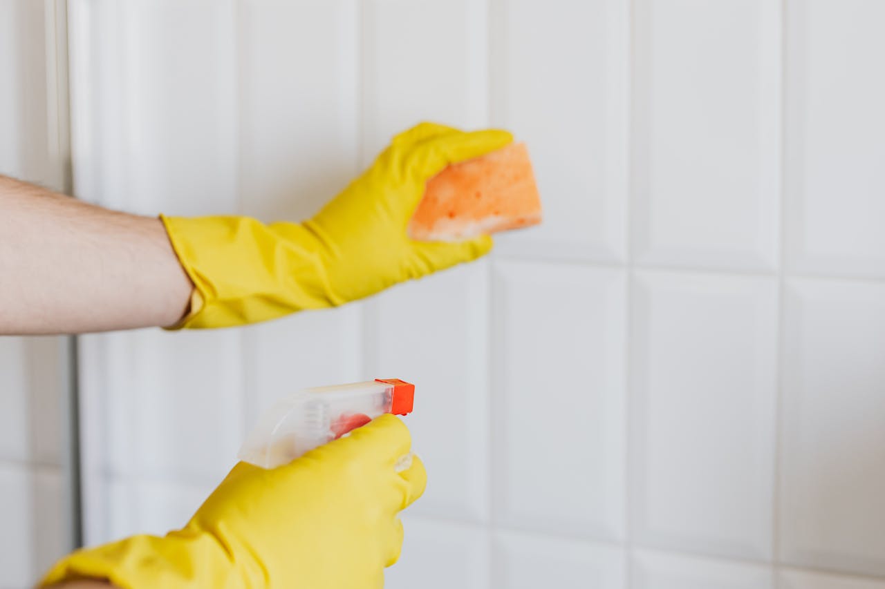 about-01 A person wearing yellow gloves cleans bathroom tiles with a sponge and spray bottle.